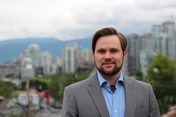 A man with a beard wearing a grey blazer and blue button-down shirt against a blurred city backdrop.