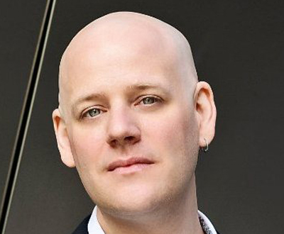 A close-up portrait of a bald man with light eyes looking toward the camera.