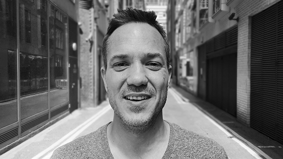 Black and white close-up portrait of a smiling man standing in a narrow urban alleyway.