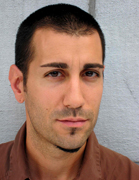 A man with short dark hair and light stubble looking directly at the camera against a grey wall.