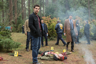 A man in a black jacket stands in a wooded area near a crime scene with evidence markers and a body.
