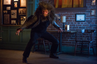 A person in a werewolf costume crouches in a dimly lit room with brick walls.