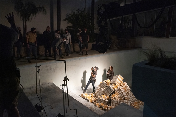 A film crew stands around a swimming pool at night, filming an actor next to a pile of wrapped packages.