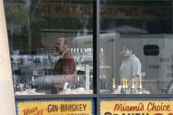 Two men stand behind a glass window filled with rows of various liquor bottles.
