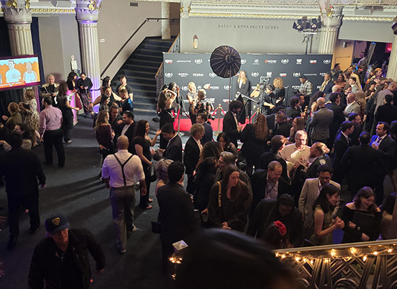 A large crowd of people gathers in a grand lobby with columns and a staircase during an evening event.