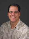 A smiling man with dark hair and glasses wearing a light-colored patterned button-down shirt.