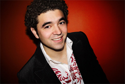 A smiling man with dark curly hair wearing a black blazer over a white patterned shirt against a red background.