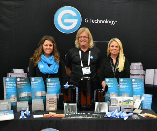 Three people pose behind a display table of G-Technology storage devices and accessories.