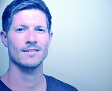 A close-up portrait of a man with short hair looking forward against a light background.