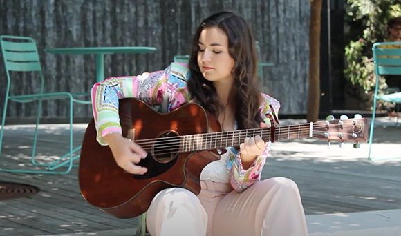 A young woman with long dark hair sits on a wooden deck playing a dark wood acoustic guitar.