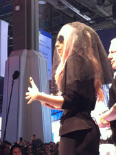 A woman with pink hair wears a black veil and outfit, gesturing with her hands on a stage in front of a crowd.