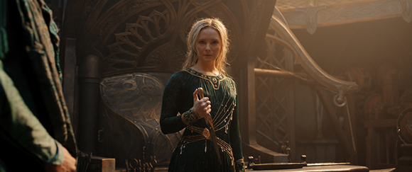 A blonde woman in an ornate dark green dress holds a dagger in a dimly lit, rustic interior.
