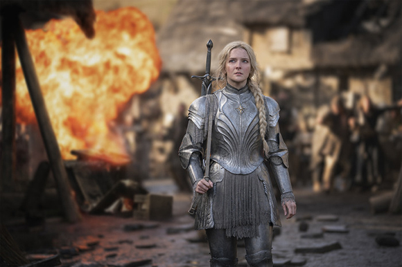 A woman in ornate silver plate armor stands in a village with a large fire burning in the background.
