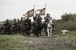 Armored knights on horseback ride across a grassy field, carrying large banners and spears.