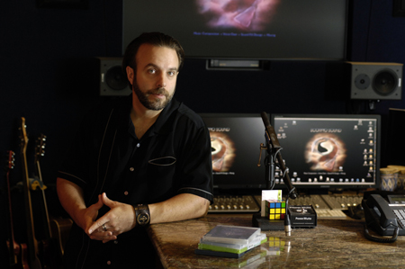 A man with a beard sits at a desk in a music studio with monitors, speakers, and instruments.