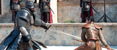 A person in full plate armor swings a longsword at a man in leather armor during a duel.