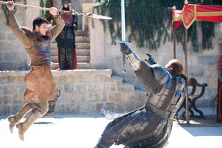 A man in brown tunic jumps with a spear while a knight in silver armor raises a sword.