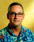 A man with short hair and glasses wearing a blue tropical print shirt against a yellow background.