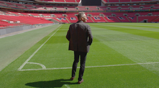 A man in a dark suit stands on a green soccer pitch looking toward empty red stadium seats.