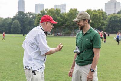 An older man in a red cap gestures while talking to a man in a green polo and tan cap in a grassy park.