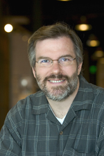 A smiling man with short brown hair, glasses, and a salt-and-pepper beard wearing a grey plaid shirt.