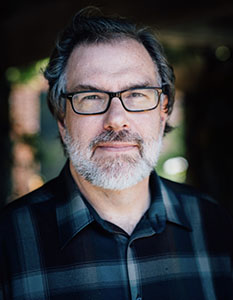 A man with salt-and-pepper hair, glasses, and a grey beard wearing a dark plaid shirt.
