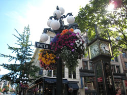 Street lamp with hanging flowers and a "WATER" sign, next to a vintage clock tower on a sunny day.
