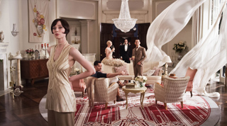 A woman in a tan dress stands in a grand room with people and flowing white fabric in the background.