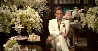 A man in a white suit sits in a room filled with white flowers and tiered dessert stands.