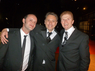 Three men in dark suits and ties smiling together at an event.