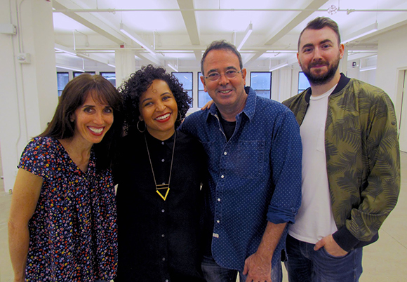 Four smiling people standing close together in a bright, white-walled room.