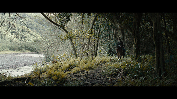 A person on a dark horse rides along a dirt path through a dense forest beside a flowing river.