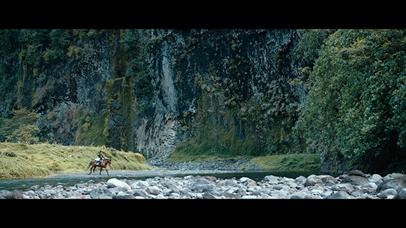 A person on horseback travels along a rocky riverbed beneath a massive, mossy cliff face.