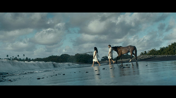 Two people in white clothing walk a brown horse along a dark sand beach under a cloudy sky.