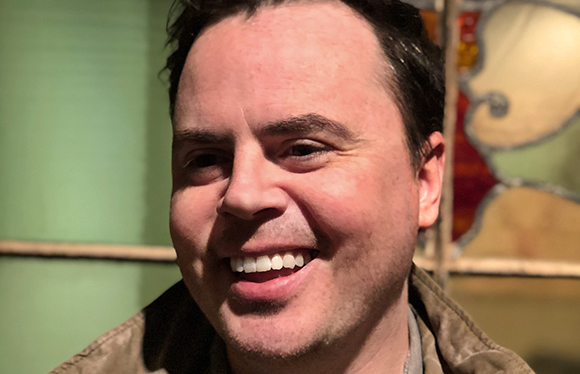 A man with dark hair and a wide smile looks toward the side.