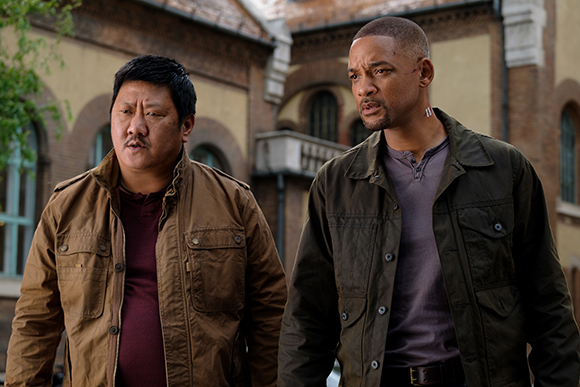 A man in a brown jacket and a man in an olive green jacket stand outdoors near a brick building.