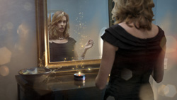 A woman in a black dress looks into a gold-framed mirror with glowing golden particles in the air.