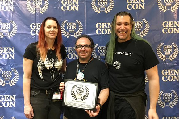 Three smiling people standing together holding a Gen Con Film Festival award in front of a blue branded backdrop.