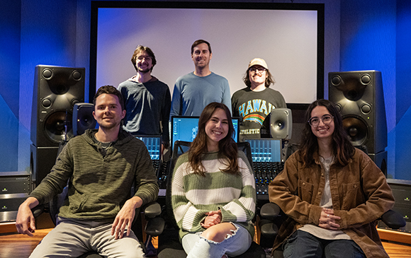 Six people pose in a recording studio with large speakers and a mixing console.