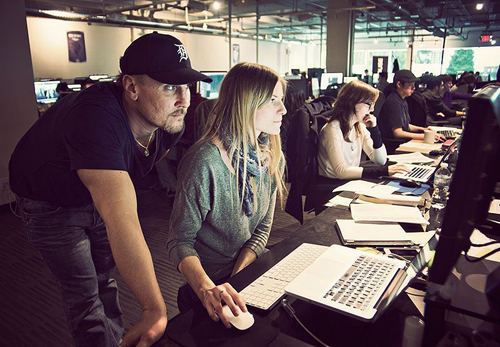 A man in a black cap leans over a woman working on a laptop in a busy, modern office.