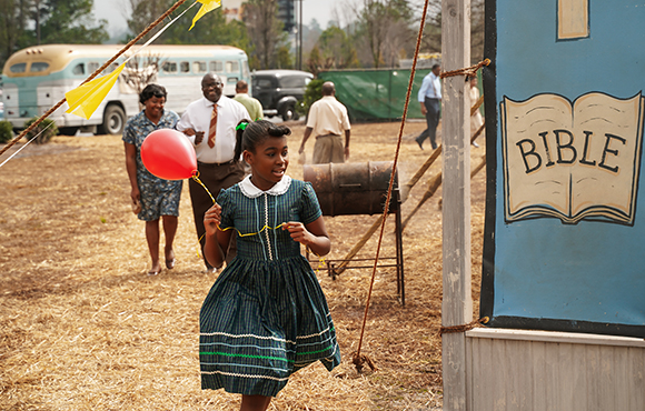 A young girl in a blue striped dress runs through a dry field past adults and a sign that says BIBLE.