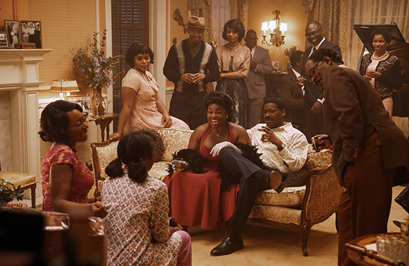 A group of Black people in vintage 1960s clothing socialize in a warmly lit, elegant living room.