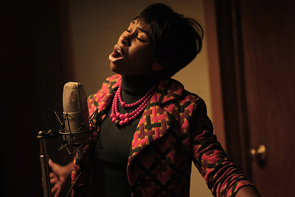A woman with short hair sings with eyes closed into a vintage studio microphone.