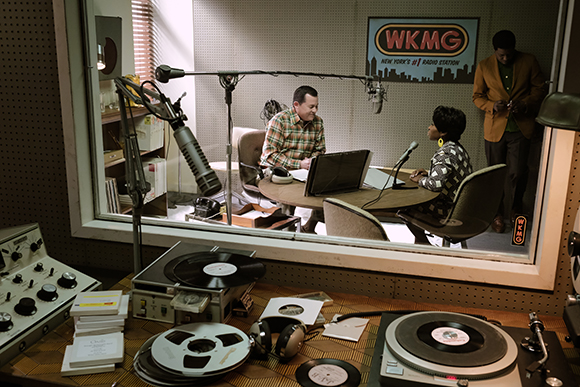 Two people sit at a desk in a radio studio booth with microphones and a sign reading WKMG.