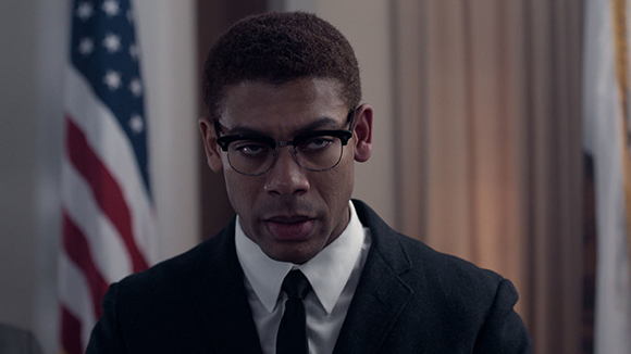 A man with short hair and glasses wearing a black suit, white shirt, and black tie.