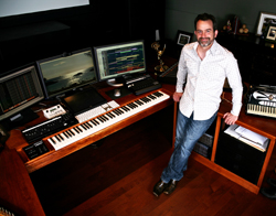 A man in a white shirt and jeans stands beside a wooden desk equipped with multiple computer monitors and keyboards.