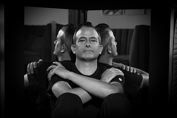 Black and white portrait of a man looking forward with his arms crossed, flanked by his side profiles in mirrors.