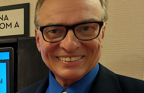 A smiling man with short hair and dark-rimmed glasses wearing a blue shirt and dark blazer.