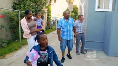 A woman carrying a child and a man in a blue plaid shirt walking toward the camera on a sidewalk.