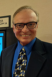 A smiling man wearing black-rimmed glasses, a blue shirt, and a patterned tie.